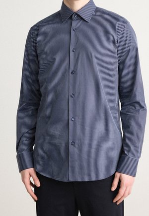 Homme portant une chemise bleu marine à manches longues avec de fines rayures verticales blanches, boutonnée, associée à un pantalon noir.