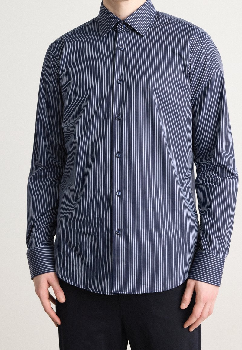 Homme portant une chemise bleu marine à manches longues avec de fines rayures verticales blanches, boutonnée, associée à un pantalon noir.