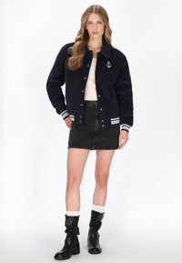 Marineblauwe corduroy varsityjack met grijze gestreepte boorden en kraag. Voorzien van een wit ankerembleem. Gecombineerd met een zwarte denimrok en zwarte laarzen.