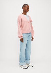 Rozā adīta jakas ar V-veida kaklu, dekorēta ar ziedu. Pāris ar gaiši ziliem platām biksēm un baltām kedām.