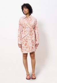 Robe chemise à manches longues en rose clair avec des motifs abstraits orange, ceinturée à la taille. Associée à des sandales à lanières rouges. Texture de tissu lisse.