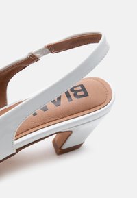 Λευκό δερμάτινο γυναικείο παπούτσι slingback με χαμηλό χοντρό τακούνι και ταμπά μαξιλαρωμένο πέλμα που φέρει μαύρο μερικό κείμενο επωνυμίας.