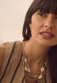 Gold-tone metal necklace with intricate leaf-shaped pendants and a double chain. Model wears a striped top, featuring a neutral color palette.