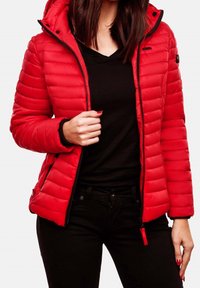 Eine Frau trägt eine rote gesteppte Zip-Jacke über einem schwarzen V-Ausschnitt-Shirt und schwarzen Hosen, mit roten lackierten Nägeln, die an ihren Händen sichtbar sind.