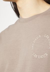 Světle hnědé bavlněné tričko s žebrovaným výstřihem a kruhovou bílou výšivkou s textem "LES DEUX" v různých umístěních.
