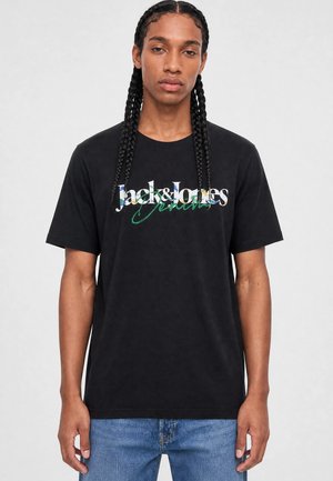 Homme aux cheveux tressés portant un t-shirt noir Jack & Jones Denim et un jean bleu, debout devant un fond clair uni.