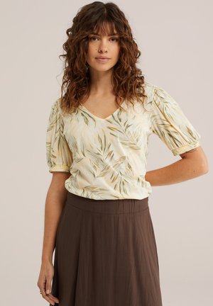 Tânără cu păr creț, purtând o bluză crem cu modele de frunze și o fustă maro cu talie înaltă, pozând pe un fundal simplu.