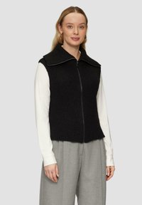 Schwarze gerippte Strickweste mit weitem Kragen und Frontreißverschluss, getragen über einem langärmeligen weißen Hemd und hellgrauen strukturierten Hosen.