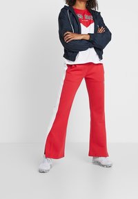 Cortavientos navy con puños elásticos, camiseta gráfica roja y pantalones rojos con rayas blancas en los lados. Zapatillas deportivas grises completan el atuendo.
