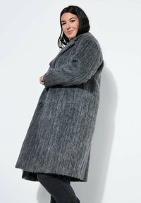 Abrigo largo gris hecho de un tejido texturizado y suave. Presenta un cuello de muesca, cierre frontal con botones y bolsillos laterales. Llevado sobre pantalones negros.