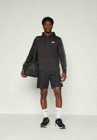 Schwarzer Hoodie mit Logo, schwarze Shorts und schwarze Sport Schuhe. Eine Umhängetasche und weiße Socken mit Logo vervollständigen den Look. Einfaches und funktionales Design.