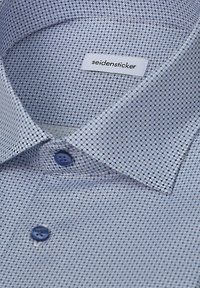 Gros plan sur le col d'une chemise à motifs avec une étiquette visible portant la mention "seidensticker" et des boutons bleus sur un tissu clair.