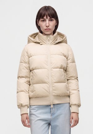 Ung person med kort brunt hår bærer en beige dynejakke med hætte og lyseblå jeans, står mod en ensfarvet hvid baggrund.