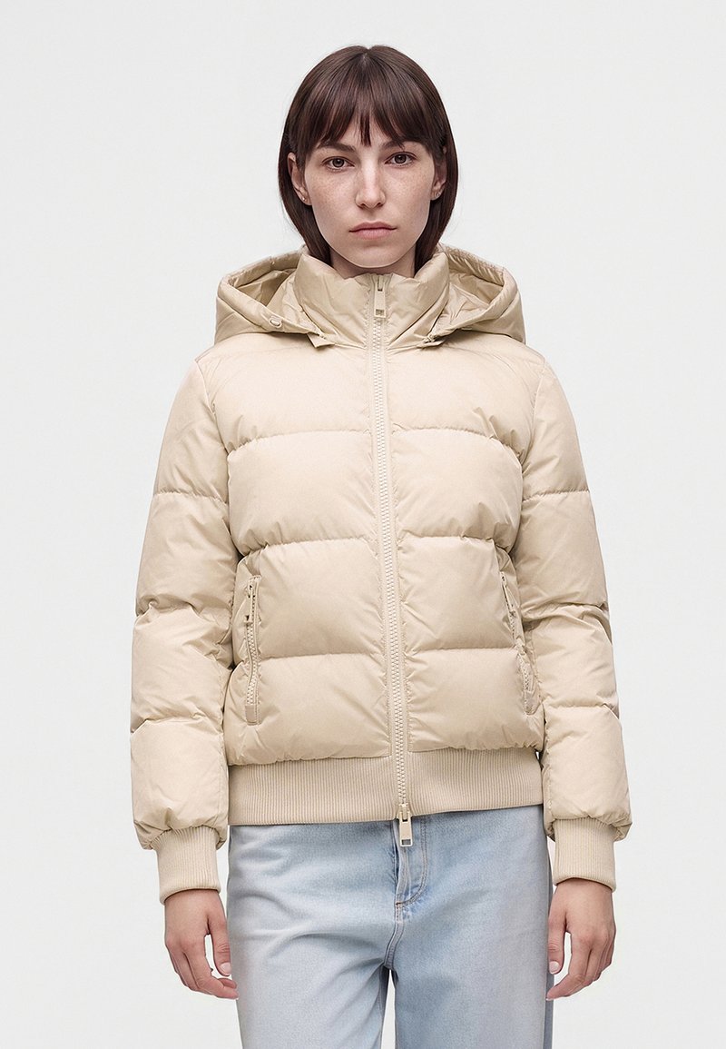 Jeune personne aux cheveux brun courts portant une doudoune beige avec capuche et un jean bleu clair, debout devant un fond blanc uni.