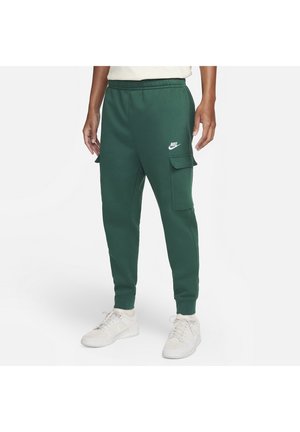 Personne portant un jogger cargo Nike vert foncé avec des poignets élastiques et des baskets blanches, debout sur un fond uni clair.