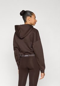 Καφέ cropped φούτερ με κουκούλα και κορδόνι, με λευκό λογότυπο, συνδυασμένο με καφέ κολάν που διαθέτουν λευκή μέση και πλαϊνές λεπτομέρειες.