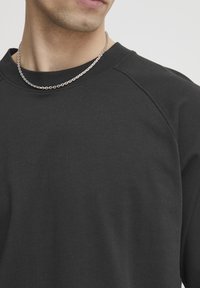 Black crew-neck sweatshirt with raglan sleeves, made from soft cotton. Features a silver chain necklace. Smooth texture, minimal design.