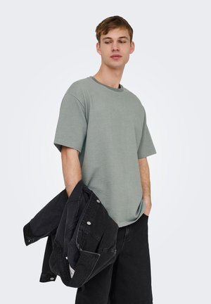 Giovane uomo con camicia verde larga a maniche corte, che tiene una giacca di jeans nera, in piedi con una mano in tasca, guarda di lato.