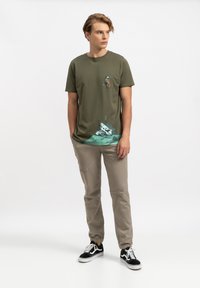 Olijfgroene t-shirt met een grafisch ontwerp van een beer en een neergestort vliegtuig, gecombineerd met beige broek en zwarte sneakers.