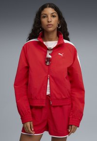 Rød letvægts zip-jakke med hvide detaljer, elastiske manchetter og en snor i kanten, parret med matchende shorts og solbriller.