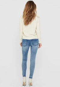Hellbeige Strickpullover kombiniert mit eng anliegenden blauen Jeans, die einen leichten Used-Look aufweisen und zwei hinteren Taschen, ergänzt durch weiße Knöchelstiefel.