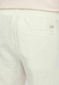 Lichtgekleurde, lichte stoffen broek met een elastische tailleband. Beschikt over een achterzak met een klein label waarop "Toegewijd aan Denim Perfectie" staat.