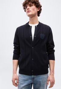 Νεαρός με σγουρά μαλλιά φοράει μια μαύρη ζακέτα με κουμπιά πάνω από ένα λευκό πουκάμισο και ανοιχτόχρωμα μπλε τζιν, στεκόμενος μπροστά σε απλό φόντο.