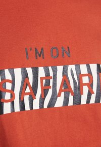 Rød bomulds T-shirt med et zebra-print stribe, der har teksten "JEG ER PÅ SAFARI" i orange og sorte bogstaver centreret på stoffet.