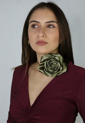 Młoda kobieta o prostych brązowych włosach nosi bordową sukienkę i naszyjnik z oliwkowozielonym kwiatem z tkaniny, patrząc lekko w lewo na jednolitym tle.