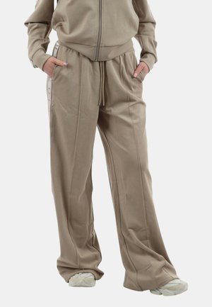 Persona che indossa pantaloni beige a gamba larga e giacca con zip abbinata, mani nelle tasche, abbinati a scarpe da ginnastica bianche su sfondo bianco.