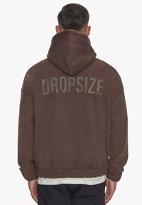 Hnedá mikina s kapucňou, vo voľnom strihu, s rebrovanými manžetami a nápisom "DROPSIZE" v kontrastnej farbe na chrbte. Mäkká textúra.