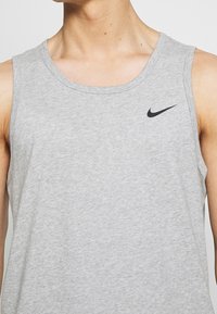 Šedý bavlněný tílko s kulatým výstřihem a černým logem Nike na levé horní straně. Měkká textura, volný design, bez rukávů.