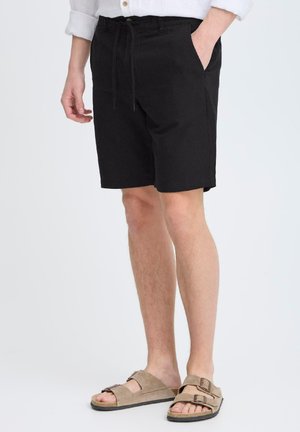 Shorts en coton noirs avec une taille à cordon, poches frontales et coupe droite, associés à des sandales beiges avec des sangles ajustables.
