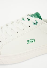White sneaker with green accents and logo. Features flat white laces, smooth leather-like material, and a textured sole for grip.