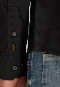 Maglietta nera con cuciture a vista, polsini a bottoni, dotata di un'etichetta con il logo di un'aquila. Abbinata a jeans in denim chiaro e sfrangiato.
