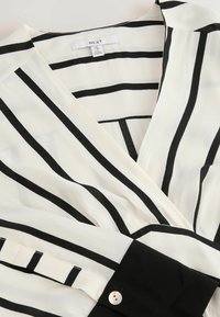 Czarna i biała bluzka w paski o kroju kopertowym, wykonana z lekkiego materiału. Posiada czarny mankiet i biały guzik przy rękawie.
