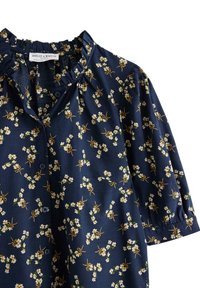 Marinebluse med blomstermønster, der har hvide blomster og gule accenter, knaplukning, rynket halsudskæring og korte, puffede ærmer.