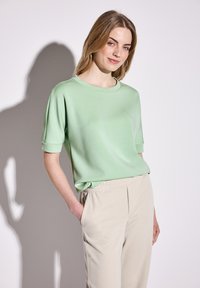 Haut vert à manches courtes avec un col rond, en tissu lisse, associé à un pantalon beige ajusté avec des poches latérales et une ceinture élastique.