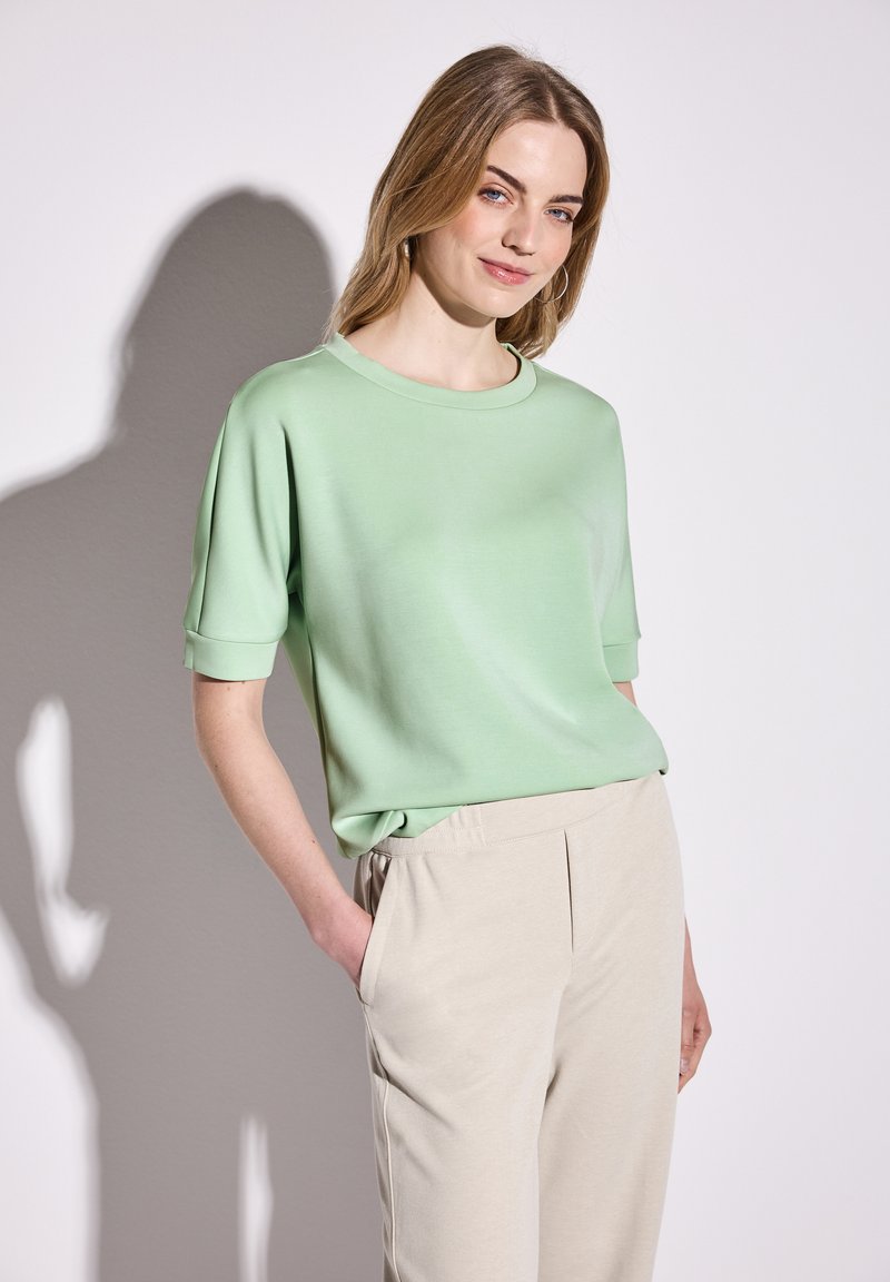 Haut vert à manches courtes avec un col rond, en tissu lisse, associé à un pantalon beige ajusté avec des poches latérales et une ceinture élastique.