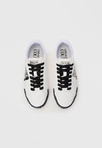 White sneakers with black laces, rubber sole, and prominent black branding on the sides. Smooth leather upper with cushioned ankle support.