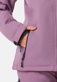 Lilla softshell-jakke med en struktureret finish, der har en lynlåslomme og et lille svensk flag-detalje på ærmet.