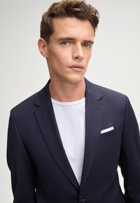 Blazer navy con revers a punta, bottone singolo e taschino con un fazzoletto bianco, indossato sopra una maglietta bianca.