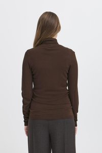 Femme aux longs cheveux bruns portant un pull à col roulé marron ajusté et un pantalon gris à motifs, debout de dos sur un fond blanc.