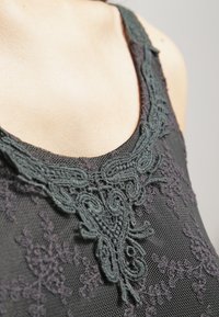 Débardeur en dentelle noire avec des détails brodés vert foncé au niveau de l'encolure, présentant un tissu en maille texturée et des motifs complexes.
