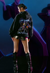 Čierna mikina s kapucňou s grafickou potlačou postáv a nápisom „GOLDEN HUNTRAX“, v kombinácii s metalickými šortkami a čiernymi čižmami po kolená.