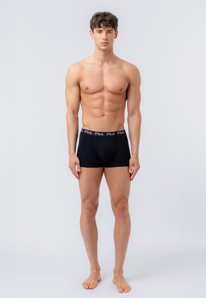 Jeune homme aux cheveux courts et bouclés, debout pieds nus, portant un boxer noir FILA sur un fond clair uni.