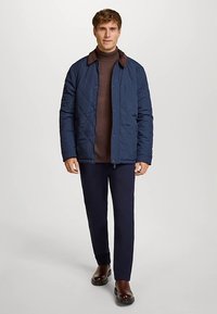 Marineblå quiltet jakke med brun krave, knaplukning og mønstret brun inderlag, kombineret med marineblå bukser og brune sko.