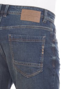Jeans de denim en azul oscuro, con una etiqueta de cuero que dice "TOM TAILOR" en la cinturilla y costuras en contraste en los bolsillos traseros.
