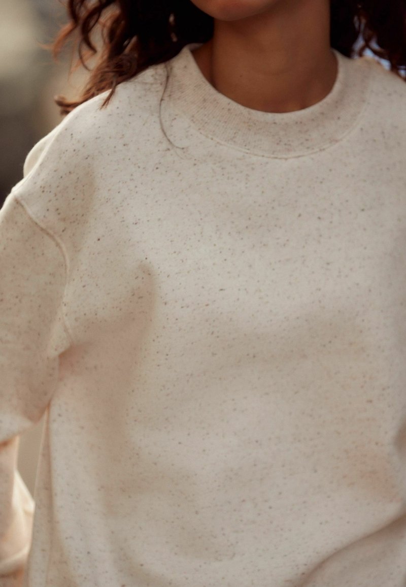 Femme portant un pull col rond beige clair tacheté à manches longues, laissant apparaître une partie de son cou et ses cheveux bouclés.