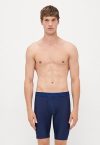 Giovane uomo con capelli corti che indossa pantaloni sportivi blu navy a compressione con elastico blu con logo, in piedi contro uno sfondo bianco uniforme.